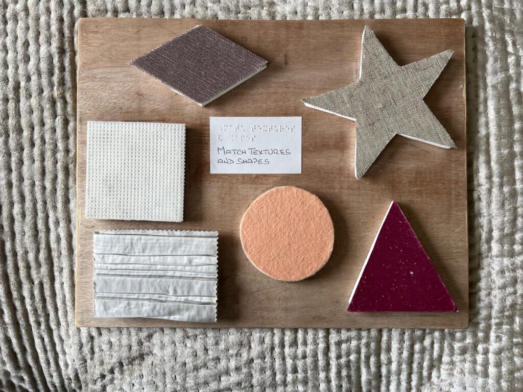 A wooden board which displays various textured shapes for tactile matching, including a star, triangle, oval, diamond, and rectangles. The center of the board contains a white rectangle card with both printed and braille text that says "MATCH TEXTURES AND SHAPES."