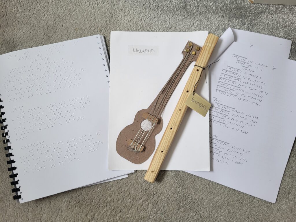 An image showing musical learning materials, including Braille documents, a handmade ukulele and a recorder, arranged on a beige carpet.