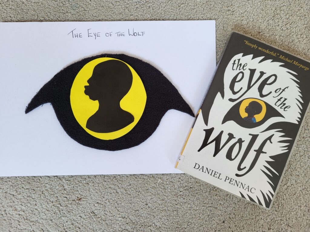 A craft project of "The Eye of the Wolf" alongside the book it's inspired by, featuring a similar design, are displayed on a carpeted surface, with the book cover quoting Michael Morpurgo.