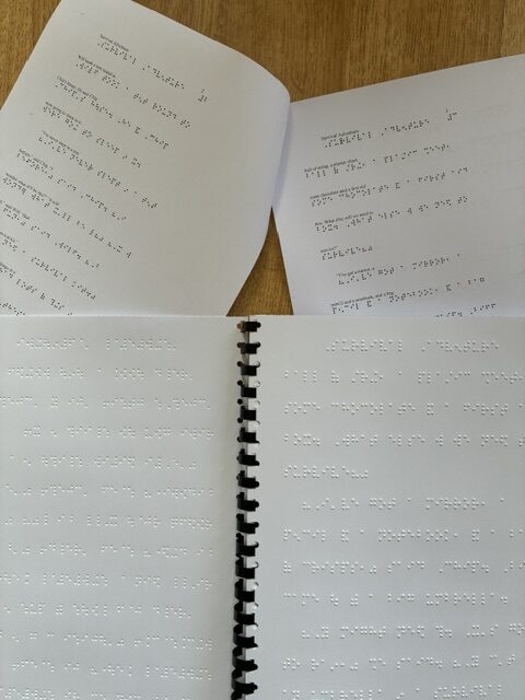 Open and spiral-bound Braille books displayed on a wooden table, showing pages with raised tactile text.