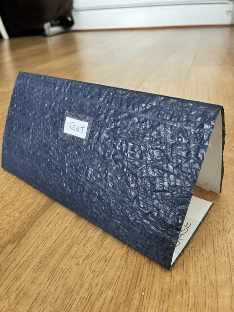 Small handmade tactile tent model made from dark-blue textured card, labeled “TENT,” standing on a wooden floor.
